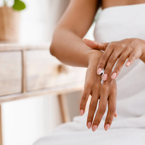 Skin nutrition concept. Unrecognizable black woman applying moisturizing cream to her hands, sitting wrapped in towel in bedroom, cropped image